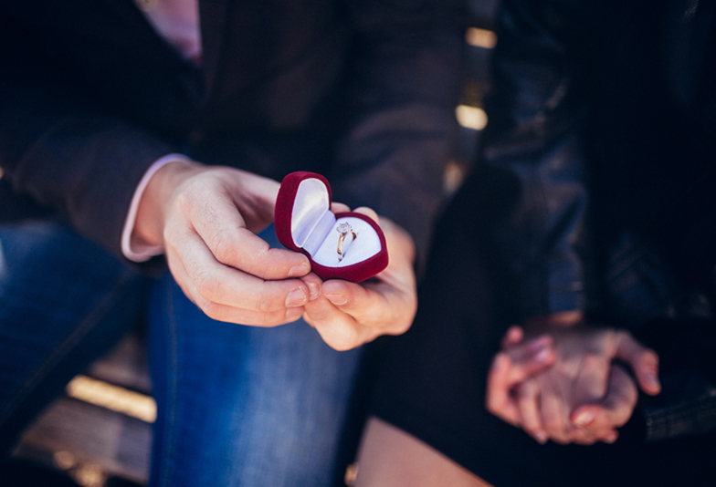 Proposal. Man's hands are holding a red gift box in the form of red heart with a ring.