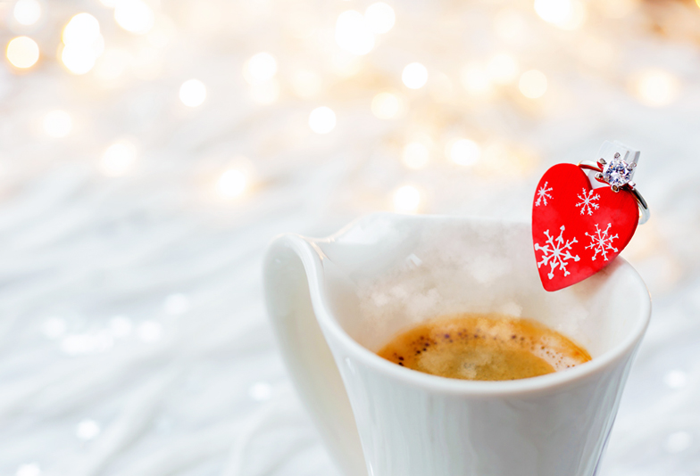 White cup of hot coffee with decorative heart and engagement diamond ring, symbol of love and marriage. Valentine's day background with lights and decorations.