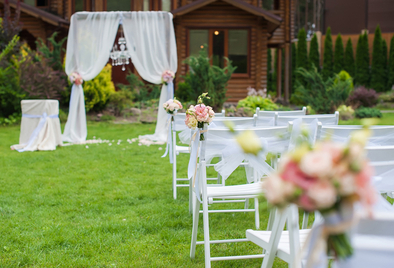 Several white chairs standing on a green lawn in front of house. White chairs decorated with bouquets of flowers and ribbons for the wedding ceremony.