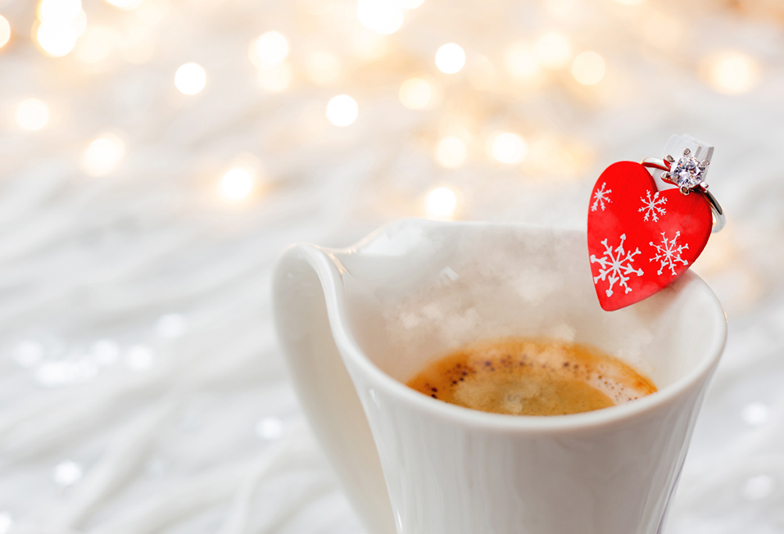 White cup of hot coffee with decorative heart and engagement diamond ring, symbol of love and marriage. Valentine's day background with lights and decorations.